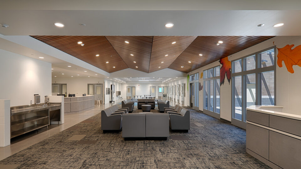 Modern waiting area with gray lounge chairs, a reception desk, large windows, and a wood-paneled ceiling.