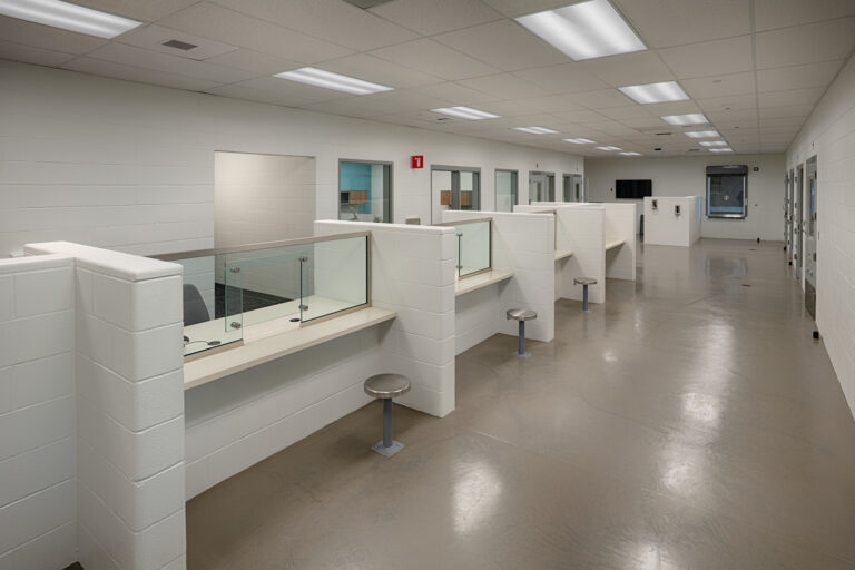 Bright institutional room with glass-partitioned interview booths, stools, and secure doors along the walls