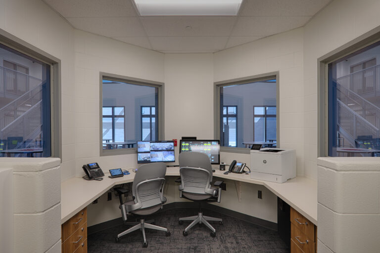 Small control room with curved desks, two office chairs, computer monitors, phones, and windows overlooking stairwells