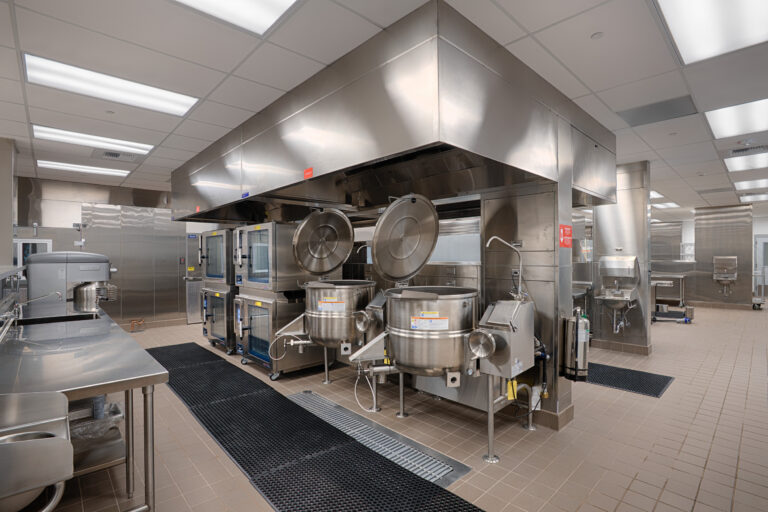 Large commercial kitchen with stainless steel kettles, ovens, prep tables, and a central ventilation hood.