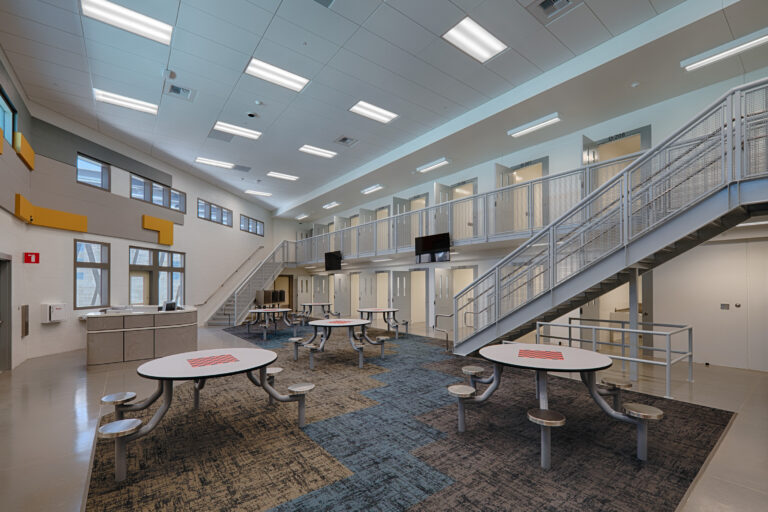 Bright two-story detention facility common area with metal staircases, upper cell doors, and round tables with fixed stools