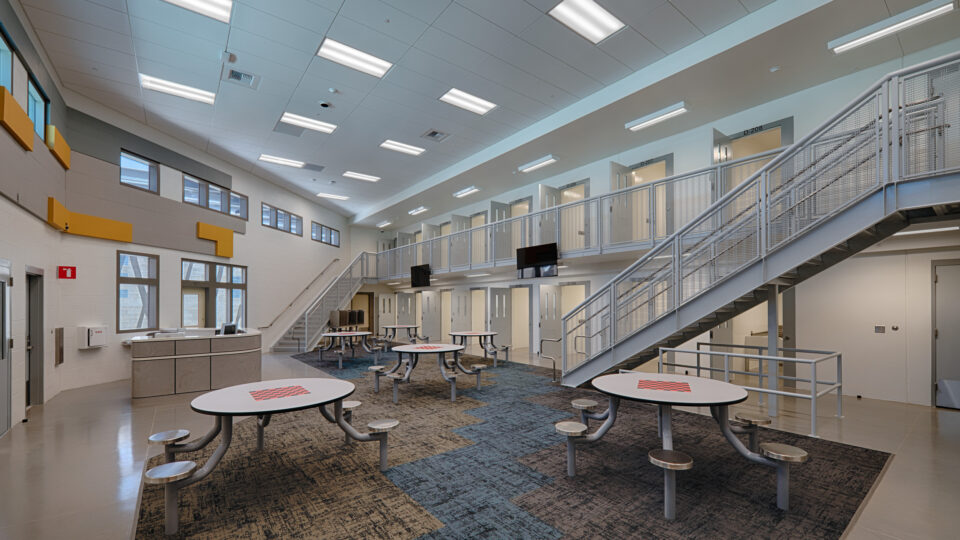 Bright two-story detention facility common area with metal staircases, upper cell doors, and round tables with fixed stools
