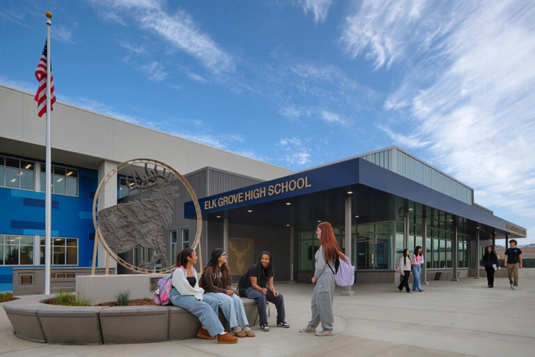 Students gather outside Elk Grove High School near a large elk sculpture and American flag under a bright blue sky.