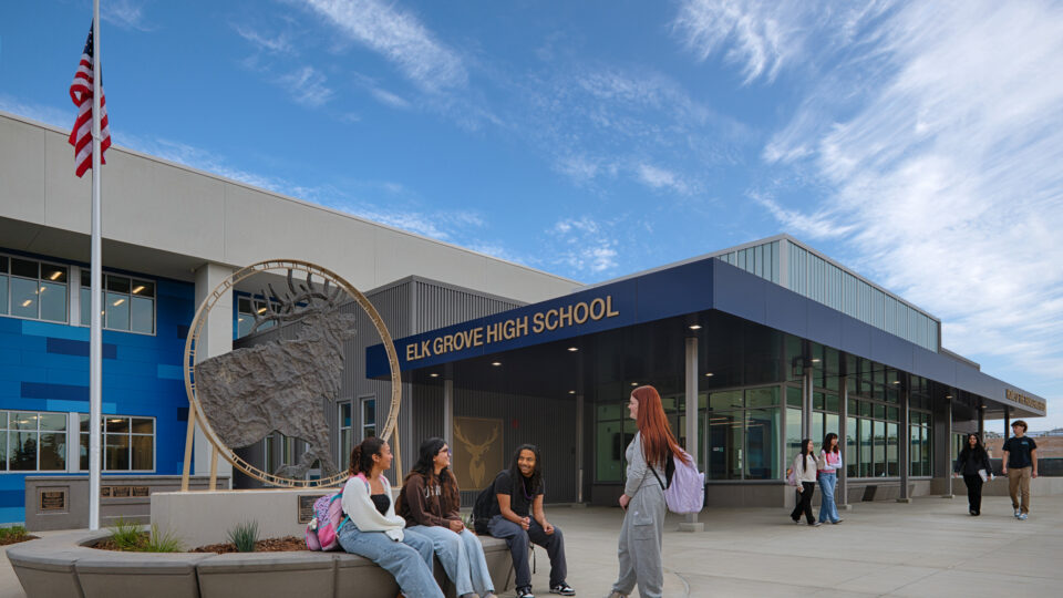 Students gather outside Elk Grove High School near a large elk sculpture and American flag under a bright blue sky.