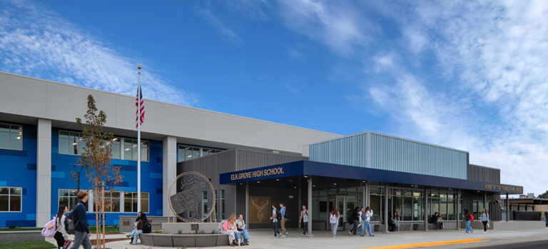 Students outside the entrance of Elk Grove High School, a modern blue-and-gray campus building under a wide cloudy sky.