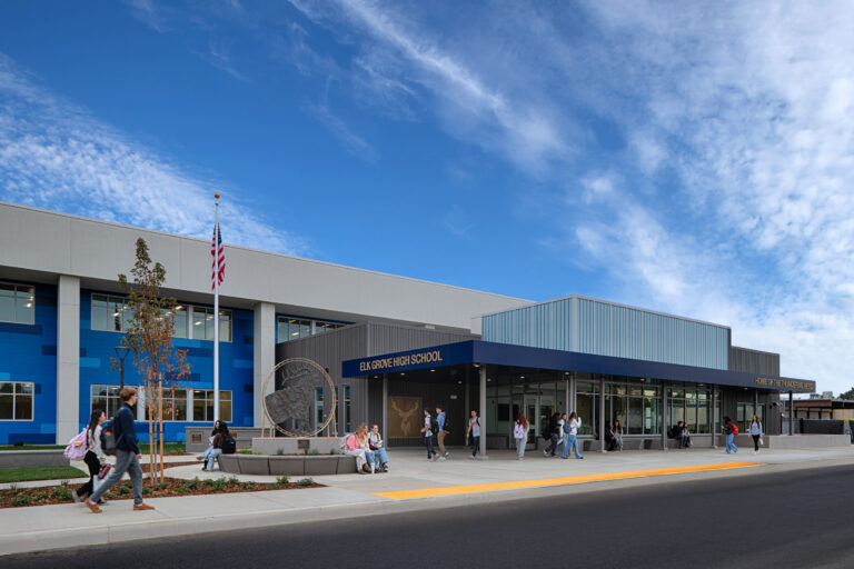 Students outside the entrance of Elk Grove High School, a modern blue-and-gray campus building under a wide cloudy sky.