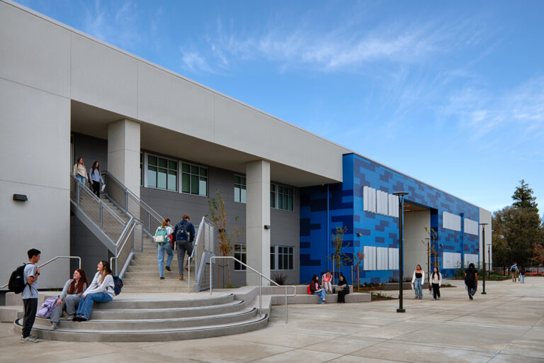 Students gather and walk outside a modern blue-and-gray campus building with wide stairs and an open courtyard.