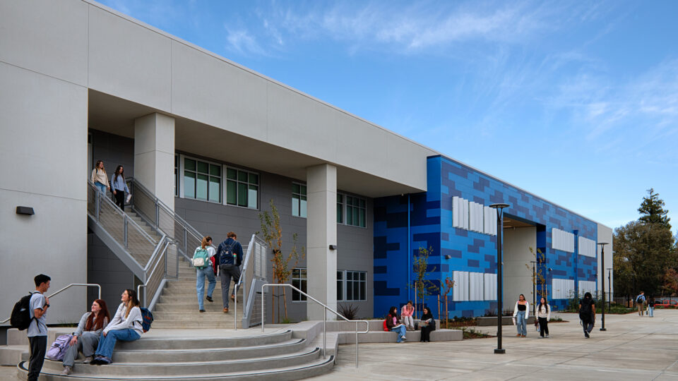 Students gather and walk outside a modern blue-and-gray campus building with wide stairs and an open courtyard.