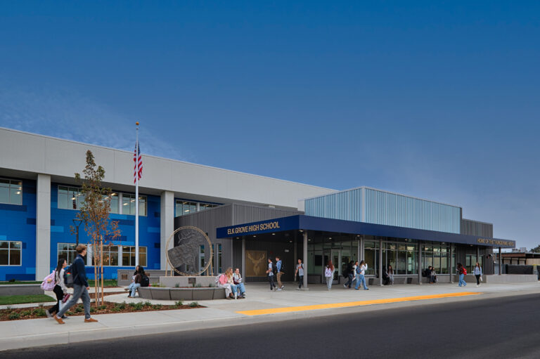 Students outside the entrance of Elk Grove High School, a modern blue-and-gray campus building with an American flag.