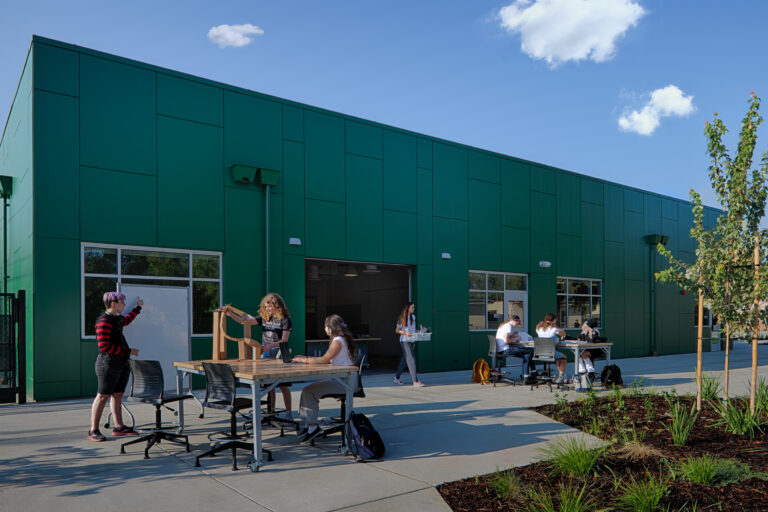 Students work and socialize at outdoor tables beside a large green building on a sunny day.