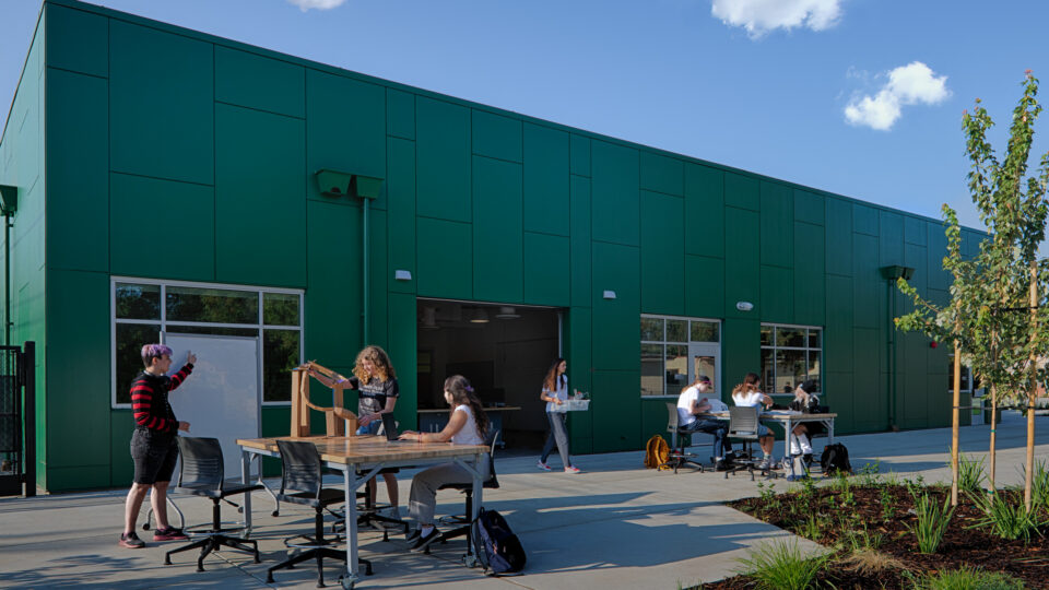 Students work and socialize at outdoor tables beside a large green building on a sunny day.