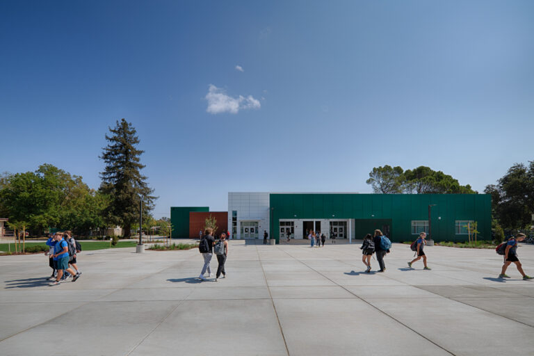 Students walking across a large plaza toward a modern green school building under a clear blue sky.