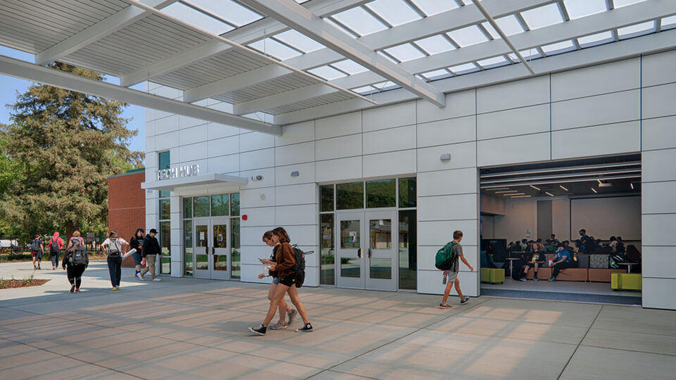 Students walk past the Tech Hub entrance beneath a large covered canopy, with more students gathered inside.