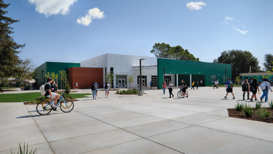 Students walk and ride bikes across a courtyard outside a modern green-and-white school building labeled Tech Hub.