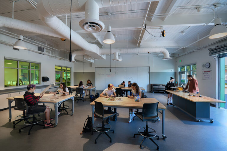 Students working at tables in a bright classroom makerspace with whiteboards, stools, and exposed ceiling ducts.