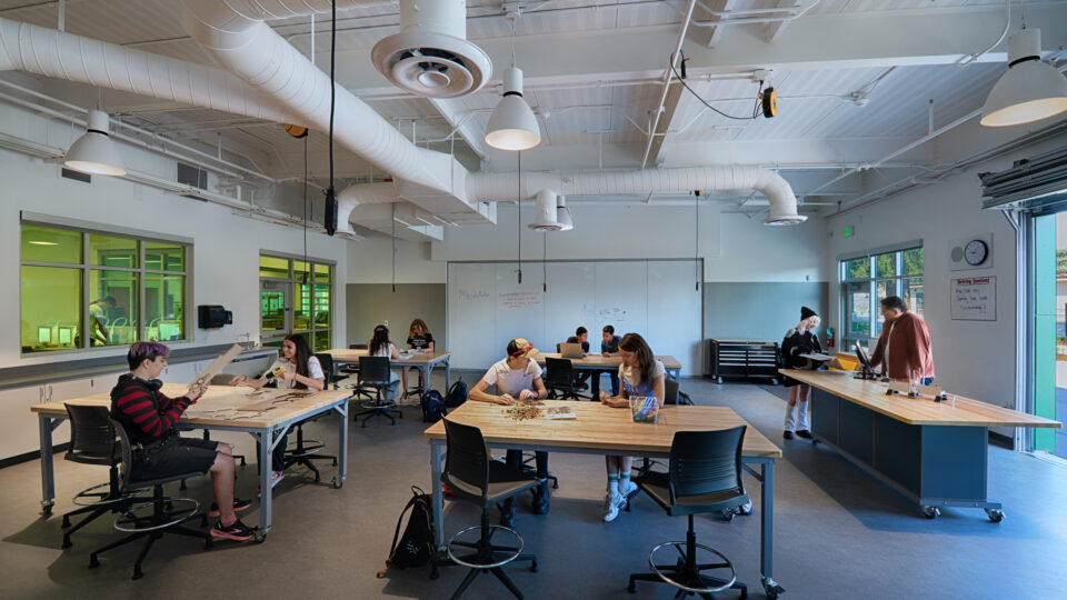 Students working at tables in a bright classroom makerspace with whiteboards, stools, and exposed ceiling ducts.