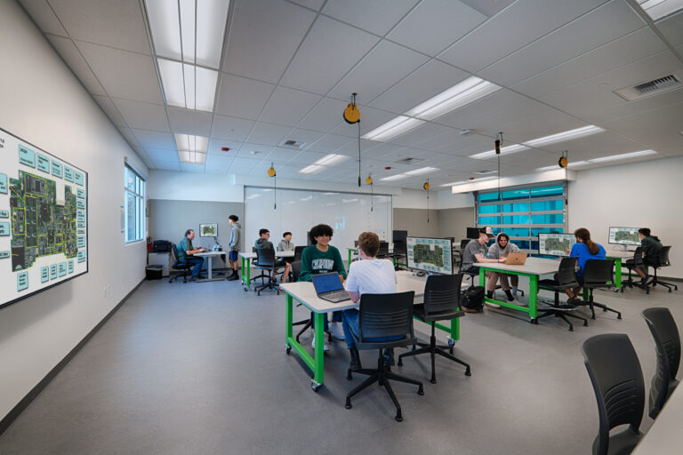 Students work at tables with laptops and monitors in a bright electronics lab, with circuit board diagrams on the walls.