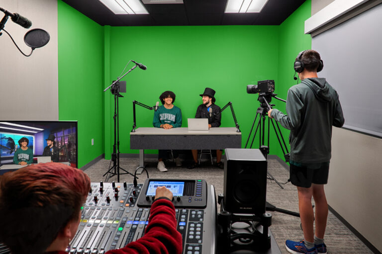 Students filming two hosts at a desk in a green-screen studio, with cameras, microphones, and audio controls visible.