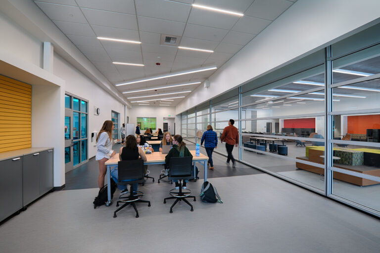 Students working and walking in a bright modern hallway with tables, glass walls, and angled ceiling lights