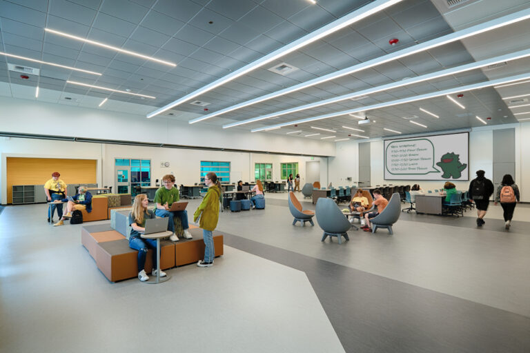 Students gather and work on laptops in a large modern school commons with tables, lounge seating, and a bell schedule screen.