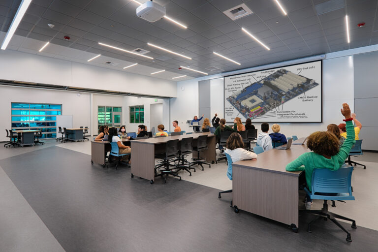 Students in a modern classroom raise their hands as an instructor presents a computer motherboard diagram on a large screen.