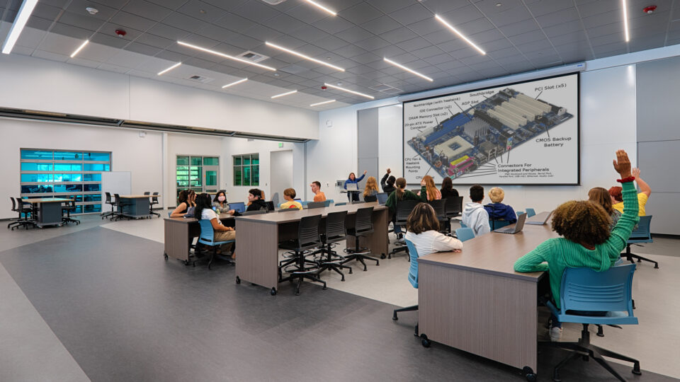 Students in a modern classroom raise their hands as an instructor presents a computer motherboard diagram on a large screen.