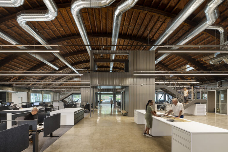 Open-plan office with exposed wood ceiling, large silver ducts, workstations, and two people talking at a counter.