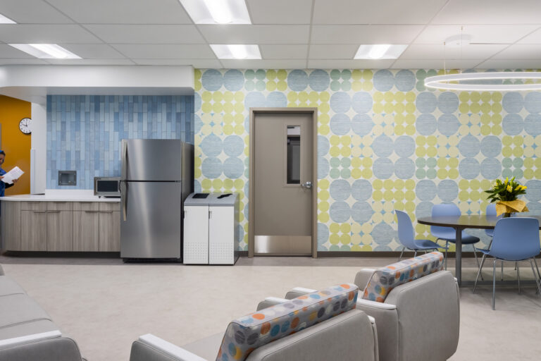 Modern staff break room with colorful patterned walls, kitchenette, lockers, seating, and two healthcare workers.