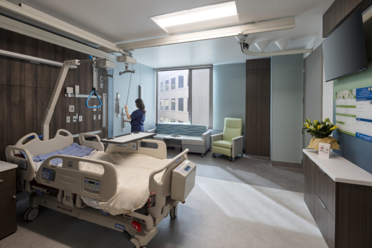 Modern hospital patient room with an adjustable bed, ceiling lift, seating area, and a nurse adjusting wall equipment.