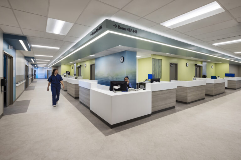 Modern hospital nurses’ station with white desks, a staff member walking, and overhead signs for rooms 425–448