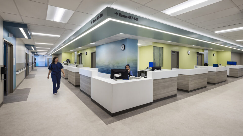 Modern hospital nurses’ station with white desks, a staff member walking, and overhead signs for rooms 425–448