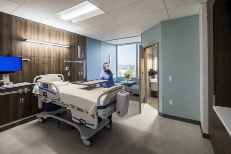Modern hospital patient room with an adjustable bed, wall-mounted monitor, bathroom, and a blurred person near the window.