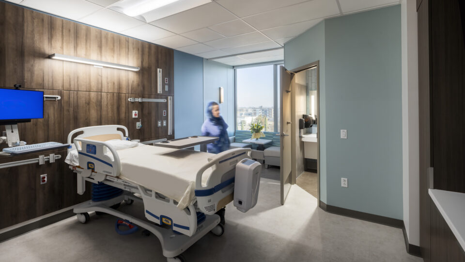 Modern hospital patient room with an adjustable bed, wall-mounted monitor, bathroom, and a blurred person near the window.