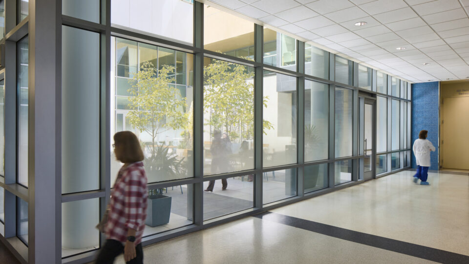 Bright hospital corridor with large glass walls overlooking an indoor courtyard, as two people walk by.