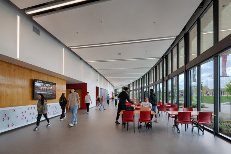 Students walking and sitting in a bright modern campus hallway with floor-to-ceiling windows and red chairs