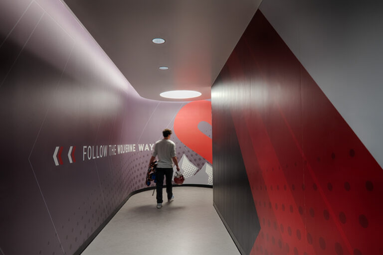 Person carrying sports gear walks down a curved hallway with wall text reading Follow the Wolverine Way.
