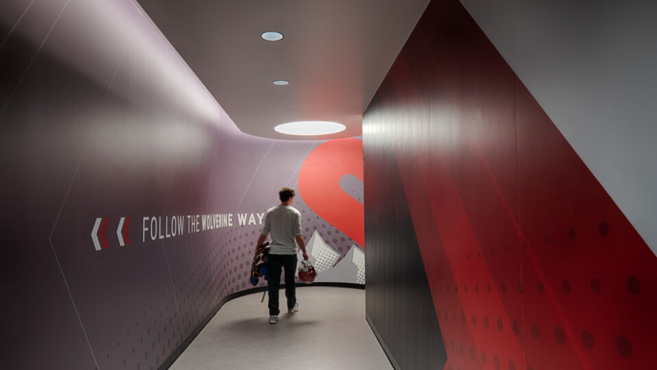 Person carrying sports gear walks down a curved hallway with wall text reading Follow the Wolverine Way.