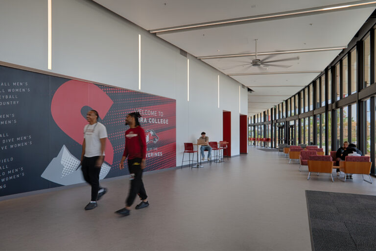 Students walk through a bright college lobby with floor-to-ceiling windows and a large Welcome to Loyola mural.