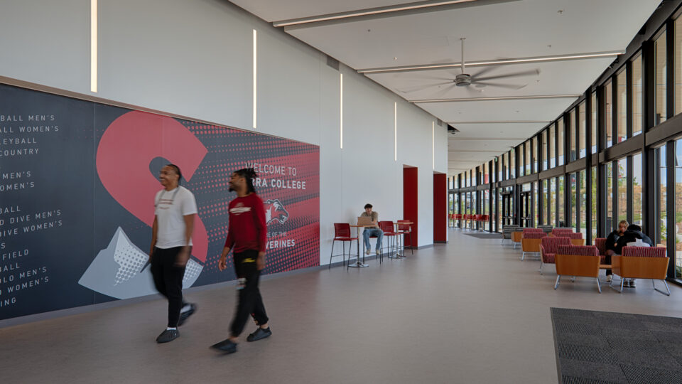 Students walk through a bright college lobby with floor-to-ceiling windows and a large Welcome to Loyola mural.