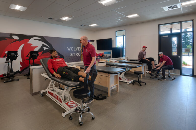 Athletes receive treatment in a university training room with exam tables, exercise bikes, and a Wolverines mural.