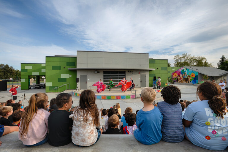 Children watch folklórico dancers perform on an outdoor stage in front of a modern green building.