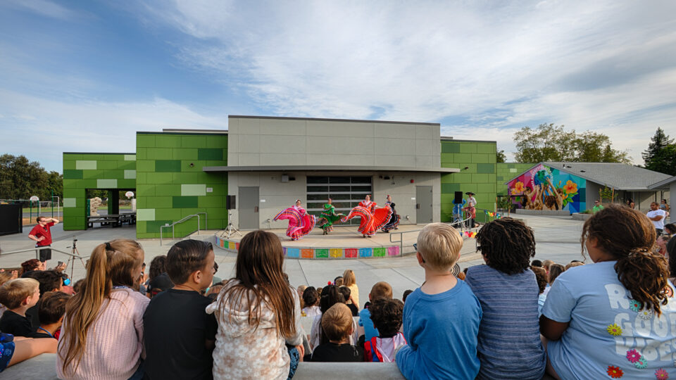 Children watch folklórico dancers perform on an outdoor stage in front of a modern green building.