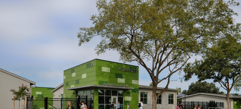 Students walking outside the green McManus Administration building beneath large trees.