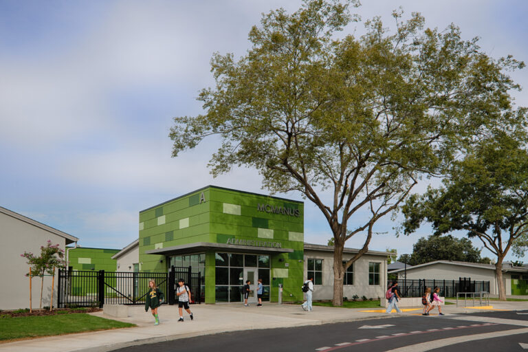 Students walking outside the green McManus Administration building beneath large trees.