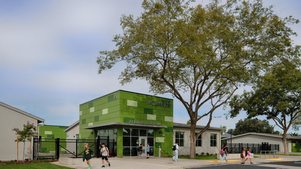 Students walking outside the green McManus Administration building beneath large trees.