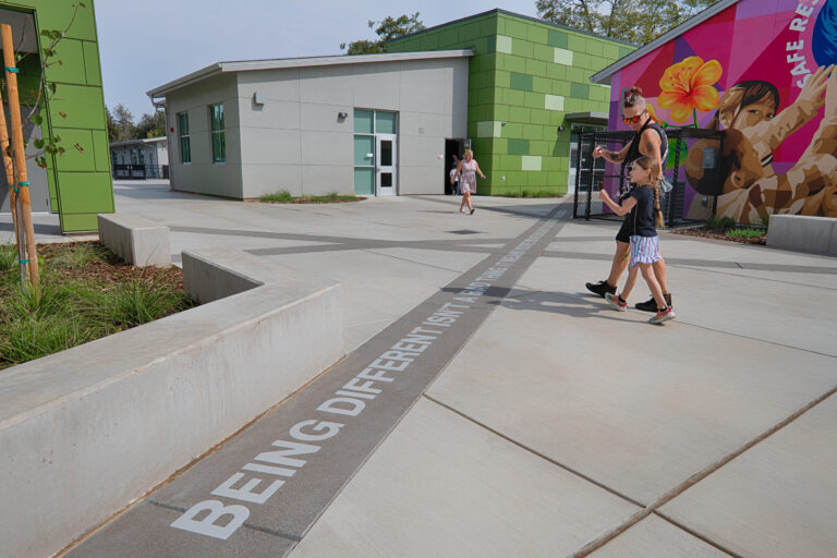 People walking through a modern courtyard between green buildings, with a colorful mural and text embedded in the pavement.