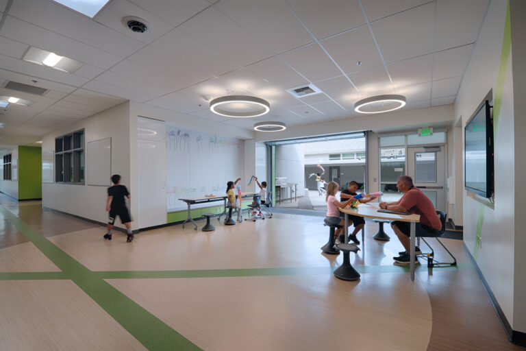 Children and an adult gather in a bright school common area with tables, stools, and circular ceiling lights.