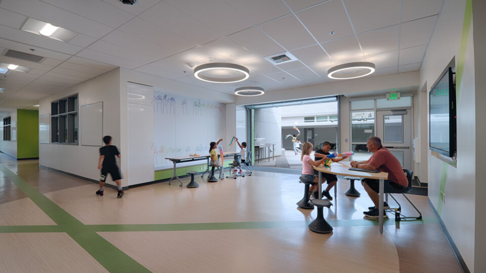 Children and an adult gather in a bright school common area with tables, stools, and circular ceiling lights.