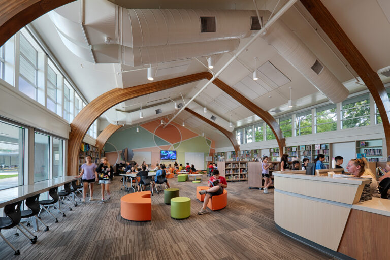 Children and adults in a bright modern library with high arched wood beams, colorful seats, and large windows.