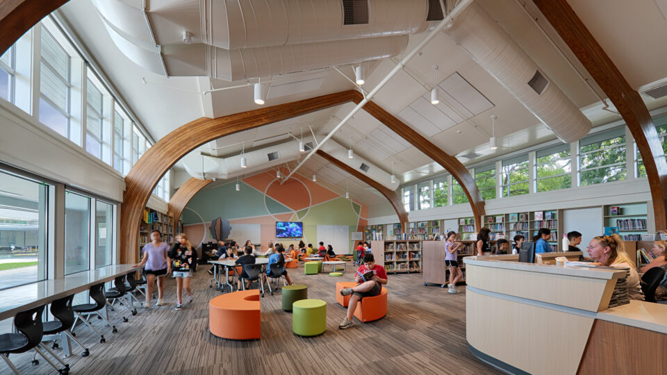 Children and adults in a bright modern library with high arched wood beams, colorful seats, and large windows.